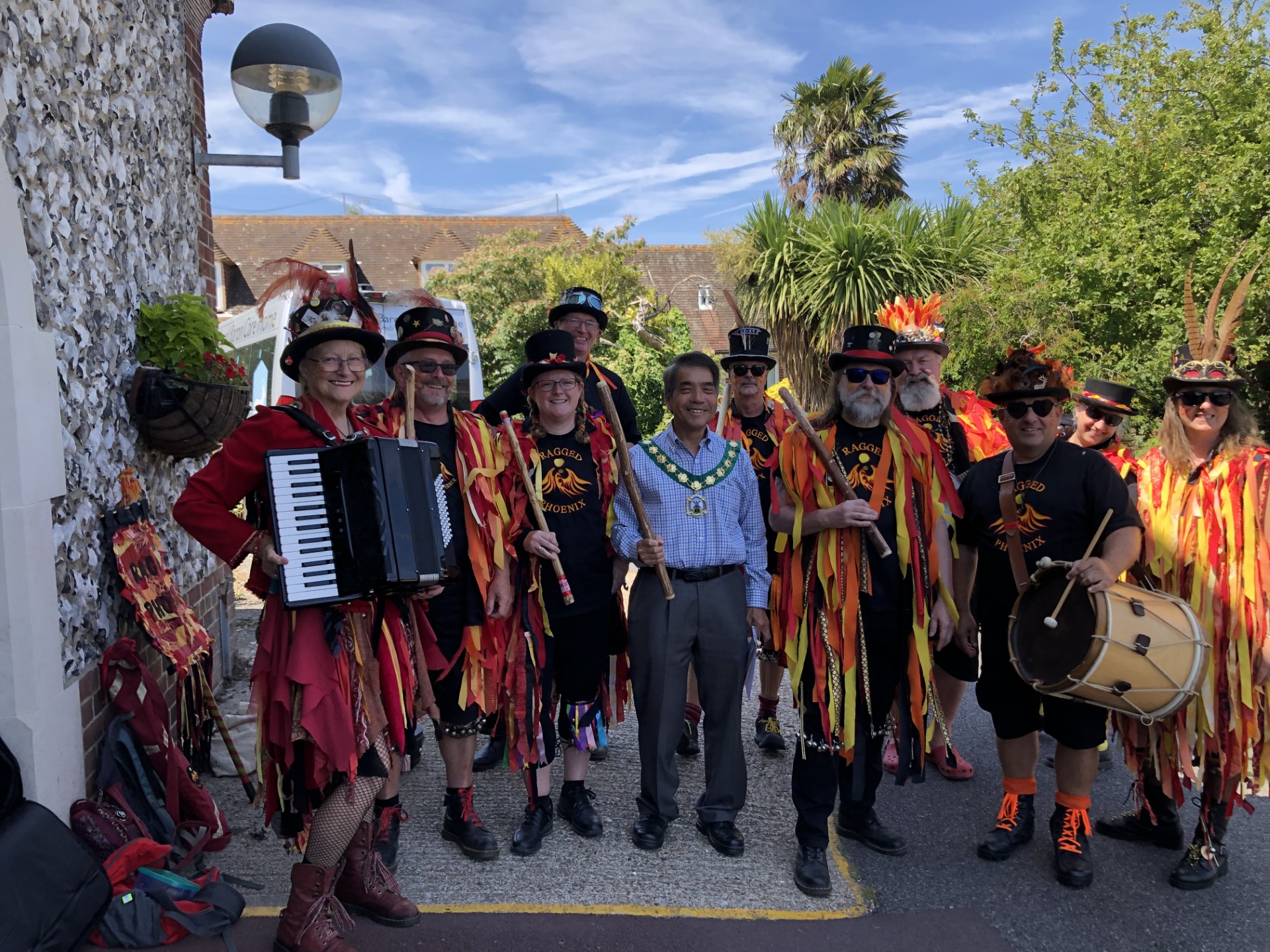 Group photograph featuring members of the Ragged Phoenix Morris Dancers in colourful red, orange and black costumes, standing outdoors with musical instruments and dance sticks at the Bernhard Baron Care Home Summer Fayre 2026.