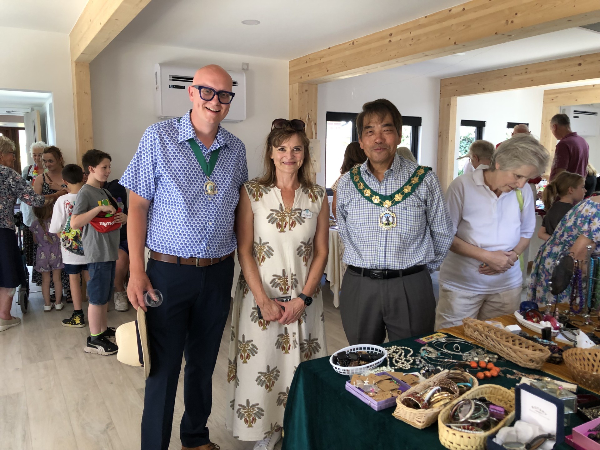 Three people standing beside a jewellery and bric-a-brac stall inside a busy community hall, with visitors browsing stalls in the background.