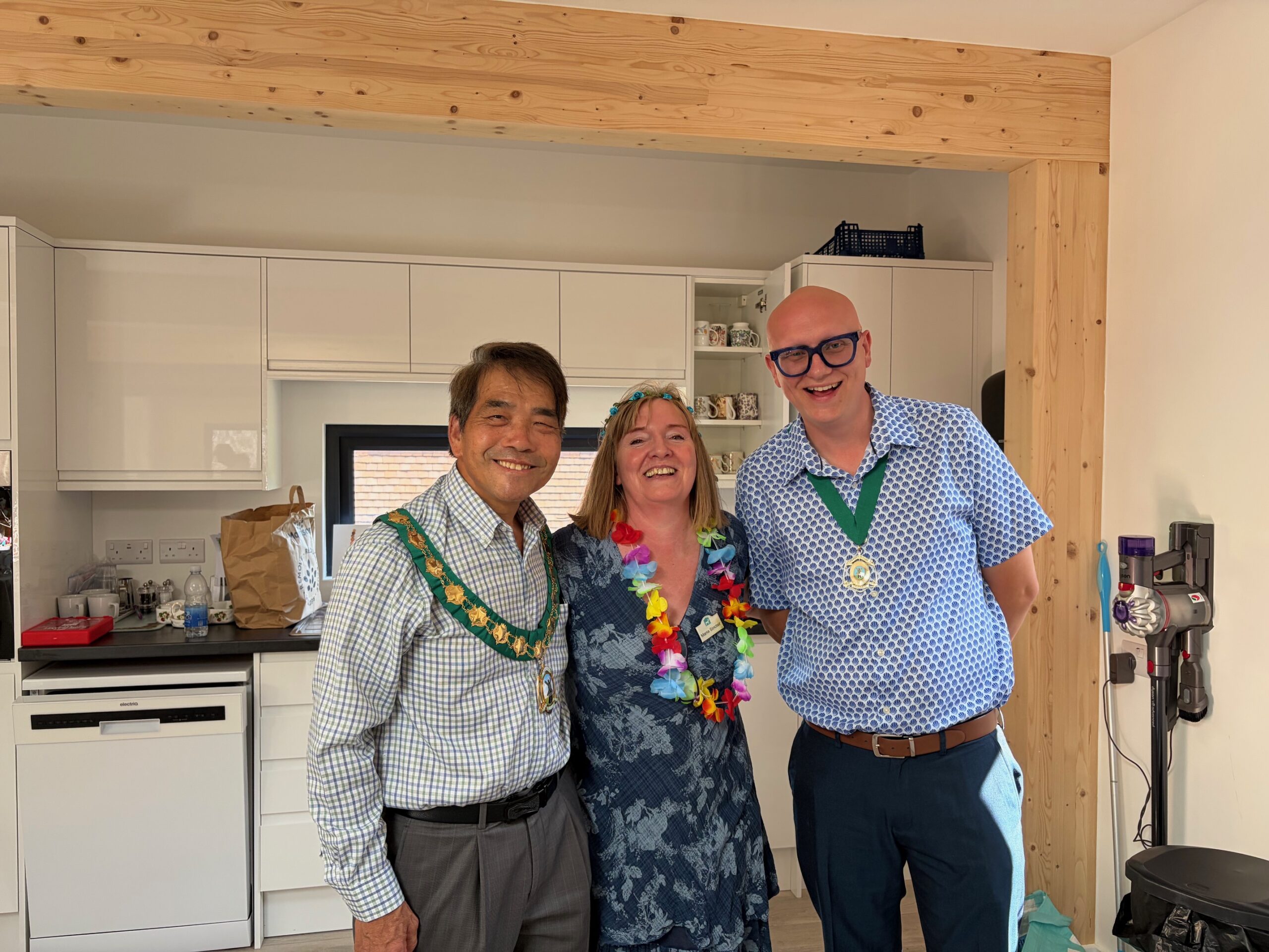 Three people standing together in a bright community room with a kitchen area in the background. One person wears a ceremonial chain and another wears a colourful flower garland.