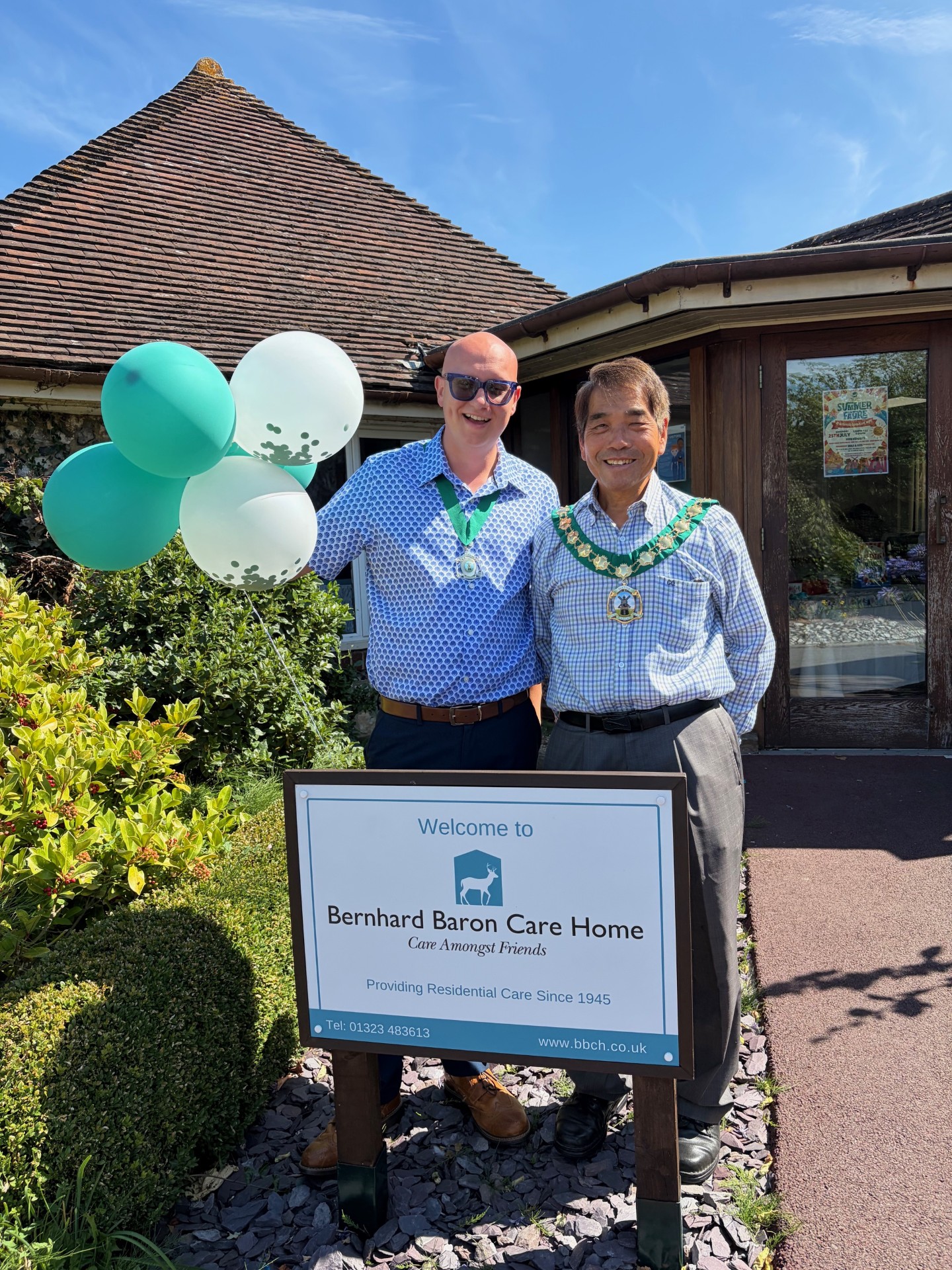 Two visitors wearing ceremonial chains stand beside the Bernhard Baron Care Home welcome sign and decorative balloons outside the care home during the Summer Fayre 2026.