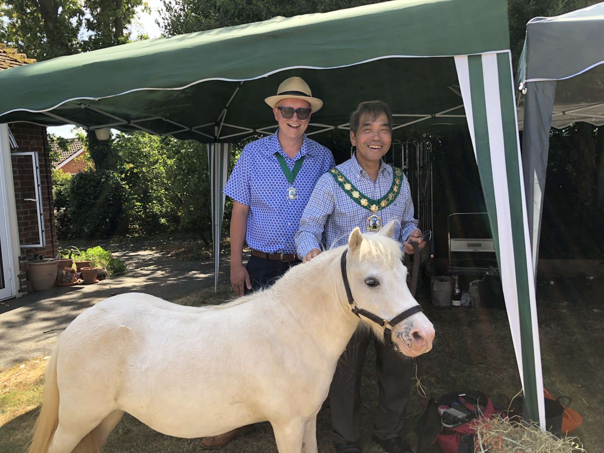 Two people wearing ceremonial chains standing beside a white pony under a gazebo at an outdoor community event.